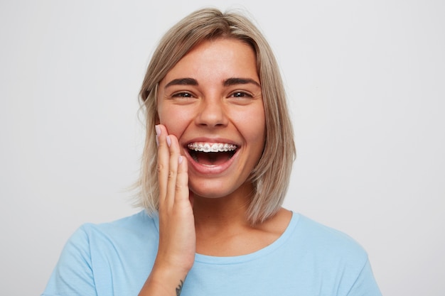 Alegre hermosa joven con cabello rubio y tirantes en los dientes riendo y tocando su rostro